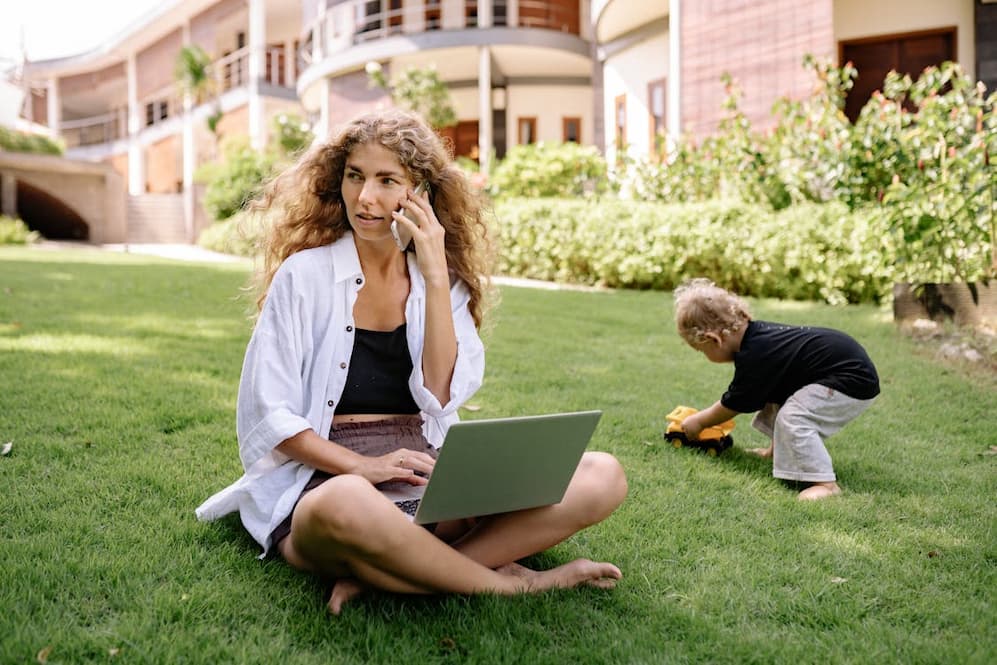 Mère en télétravail sur un ordinateur portable, son fils jouant à côté dans un parc